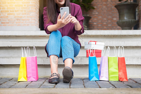 妇女看手机时在购物中心用纸袋看手机同时享受白天购物图片