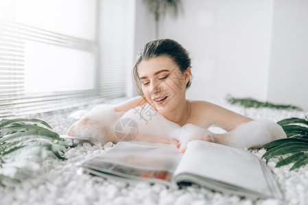 坐在浴缸里带着泡沫的微笑女士坐在浴缸里看杂志豪华浴室装有窗子和棕榈树枝装饰图片