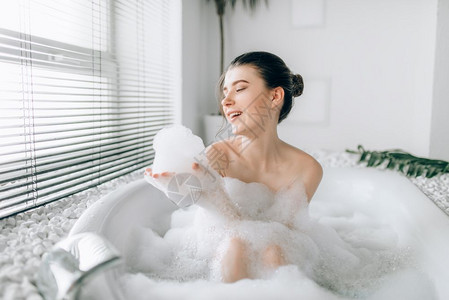 坐在浴缸里玩泡沫的笑着女人豪华的浴室有窗户和棕榈树枝装饰背景图片