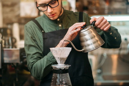 坐在黑围裙和眼镜的侍者倒在咖啡壶里的一个热煮水侍者倒在咖啡壶里热煮水图片