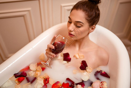 美丽的女人在带着玫瑰花瓣和泡沫浴缸里喝红酒图片