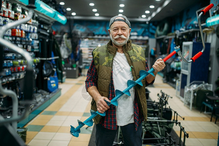 男钓鱼者购买渔获和狩猎的设备工具在商店的展品上进行各种示渔民在商店进行冬季捕鱼的钻头图片