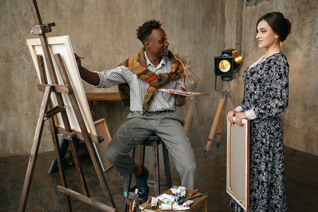 女人看着画家用调色板用画笔作画男人坐在画架前背景是艺术工作室内部创意大师在工作室一个女人看着画家用画笔作画图片