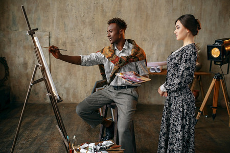 女人看着画家用调色板用画笔作画男人坐在画架前背景是艺术工作室内部创意大师在工作室一个女人看着画家用画笔作画图片