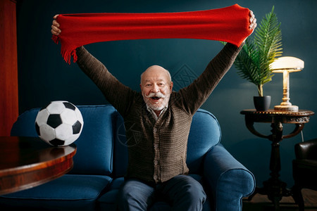 有红围巾和球观电视的老人足球迷在客厅留胡子的成熟长姿势老年人闲时间图片
