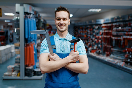 男工穿着制服微笑的男工在具商店中握着锤子和角落选择硬件商店仪器超市的专业设备工人在具商店中握着锤子和角落图片