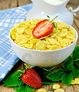 白碗中的玉米片叶子和草莓罐头中的牛奶木板背景上的餐巾纸图片
