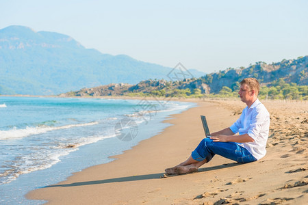 坐在沙滩上拿着笔记本电脑的英俊年轻男子图片