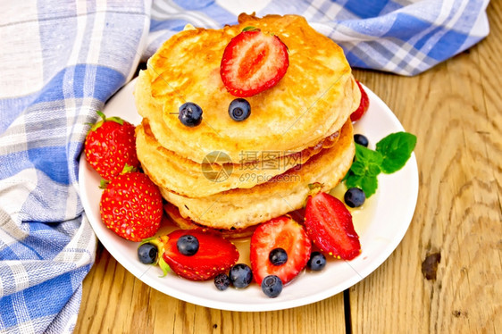 一堆煎饼上面有草莓蓝和蜂蜜放在白色盘子上餐巾纸放在木板背景上图片