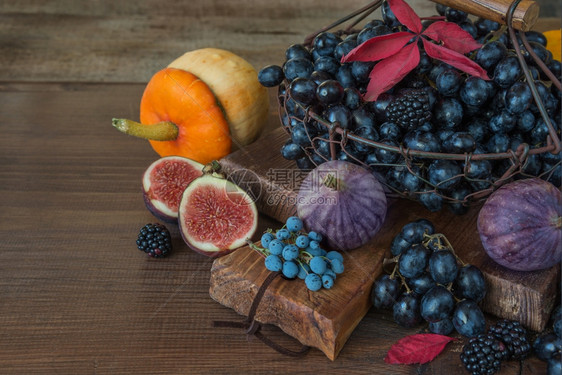 旧切割板上篮子中的黑葡萄以及成熟的无花果红草莓橙色南瓜和木本底有秋叶的黑莓图片