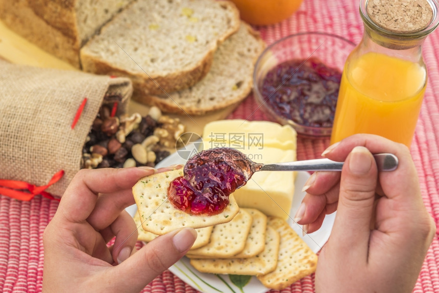 女人手握着一个装草莓果酱的饼干而在背景里早餐桌还有面包黄油橙汁和混合坚果图片