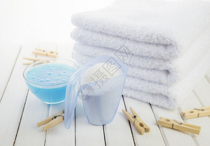 素色毛巾3条白色毛浴巾测量杯中的洗衣粉透明玻璃碗中的蓝织布软体和白板底的木制衣背景