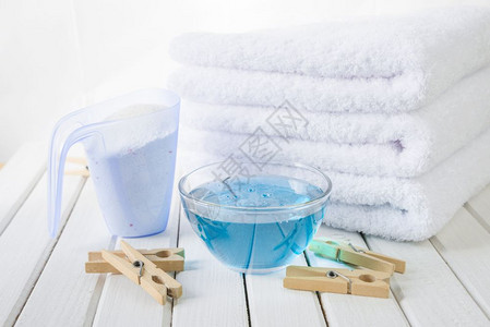 3条白色毛浴巾测量杯中的洗衣粉玻璃碗中的蓝织布软体和白板背景上的木织物图片