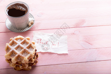 美味的自制华夫饼里面有一杯热咖啡和心形糖放在粉红色的木制桌子上还有一张纸上面写着感谢信图片