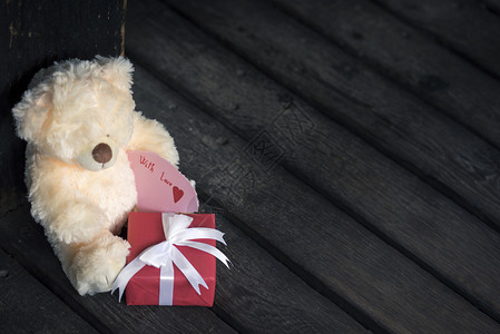 红礼盒上面有白绑的弓和可爱泰迪熊玩具在生锈的木地板上挂有口信图片
