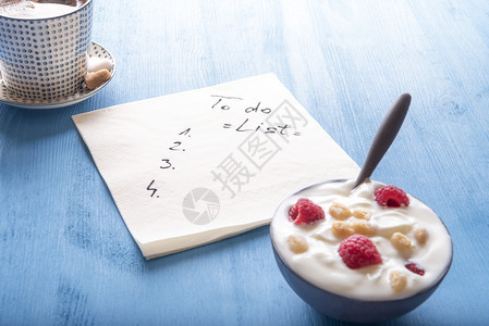 一杯阿拉伯咖啡一碗酸奶配有草莓和麦片的酸奶以及一张餐巾纸上面有一份工作清单放在蓝色木制桌子上图片
