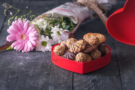 红色的心形盒子里面装满了糖巧克力饼干和一束鲜花在背景里黑木制桌子上图片