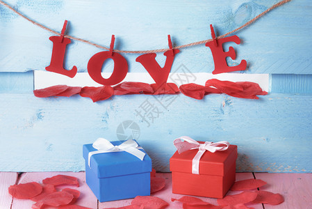被肥皂红色花瓣包围的礼物盒和用红纸信拼写的爱字挂在一根绳子上蓝木栅栏图片