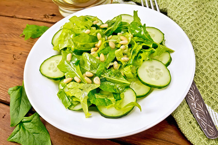 菠菜沙拉新鲜黄瓜Rukola沙拉雪松坚果和绿洋葱盘子里有植物油餐巾和木板背景的叉子图片