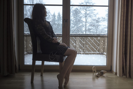 穿着黑裙子的优雅年轻黑发女人坐在椅子上放松看着大玻璃门的雪花背景图片
