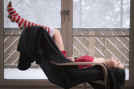穿着短红色裙子和长袜的年轻感女人坐在椅子上倒着放松站在玻璃门前外面在下雪图片
