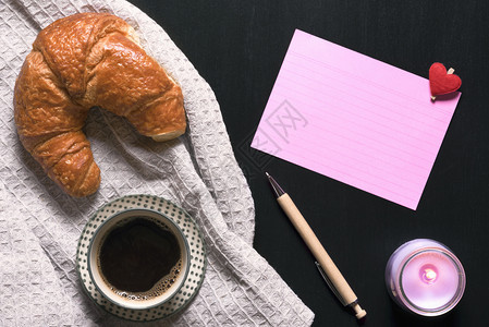 黑咖啡和羊角面包放在古老的厨房毛巾上有粉红色笔记本和黑桌上有点亮的粉色蜡烛图片
