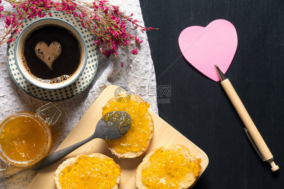 浪漫的早餐鲜花心形粘的纸条果酱在战壕的面包咖啡杯在黑桌子上图片