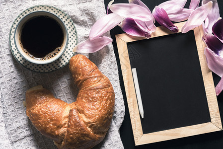 一杯热咖啡和块新鲜的羊角面包放在一块旧厨房毛巾上靠近一张空白的黑板四周是粉红色的郁金香花瓣在一张黑桌子上图片