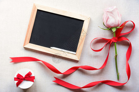 粉红玫瑰上绑着丝带和弓靠近一个可爱的白色礼品盒和一张空白的黑板以旧布料背景图片