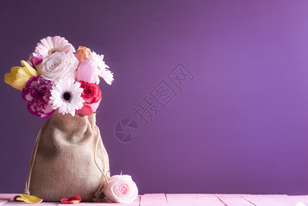 挂着花朵的玫瑰郁金香和雪贝拉的生日卡包裹在陈年的帆布袋里放在粉红色的木桌和紫背景上图片