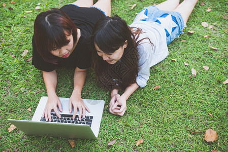 躺在草地上使用笔记本电脑和打字的年轻女孩手握键盘远程学习概念在公园里工作笔记本电脑上工作的快乐年轻女在户外学习的生图片