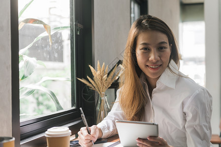 身着白衣的年轻女商人坐在咖啡厅的桌子上一边用笔记本写作一边用平板在线学习图片
