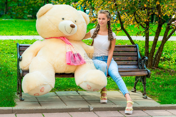 一个快乐女孩坐在长椅上与你最喜欢的泰迪熊在公园肖像图片