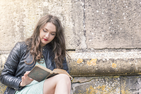 年轻女子在户外阅读一本旧书仰着石墙在阳光明媚的一天图片