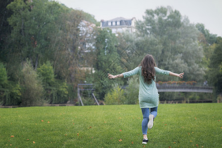 褐色的年轻女子穿着随意服装在雨中快乐地奔跑她的双臂宽阔周围是绿草和树木在德国图片