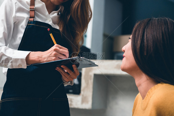 年轻的女咖啡店主在小里笑容热烈欢迎地接受客户的订单年轻女从客户那里得到咖啡食物和饮料图片