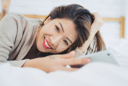 快乐的亚洲妇女早上在床使用智能手机图片