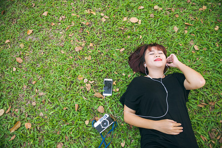 年轻女士躺在绿草地上听音乐年轻女士躺在绿草地上听公园里的音乐带着寒冷的情绪年轻女士用她的音乐播放列表在草地上松公园概念中的户外活图片