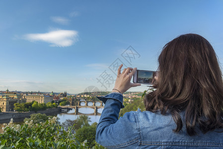 黑发女人从后面用她的智能手机拍照包括伏尔塔瓦河桥梁普莱格城市切图片