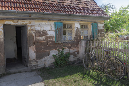 在阳光明媚的一天老化德国人房屋有剥皮的墙壁窗户上木帘开门栅栏和花园上的生锈自行车图片