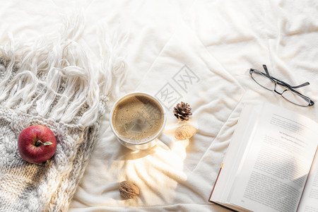 舒适的休闲概念开放的书眼镜苹果在毛披肩和热咖啡在白毯上早晨的阳光下图片