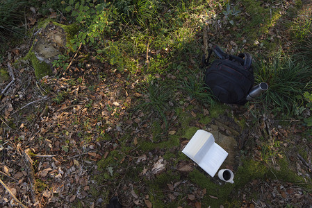 在树荫下的根上在秋天的森林地板上看到一个挂着热水瓶的徒步背包以及一本开阔的书和杯咖啡图片