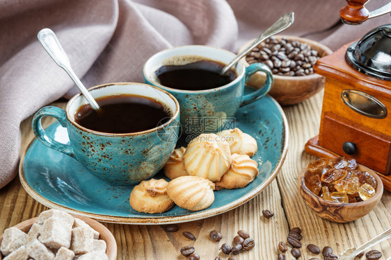 两杯黑色咖啡饼干和糖的蓝色杯子在旧木制桌布和咖啡豆上有两杯黑咖啡饼干和糖片上面有格式床单布和咖啡豆图片