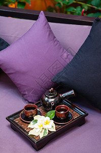 木托盘上的茶杯和手工陶瓷锅沙发上装有彩色织布枕头的亚洲式茶图片