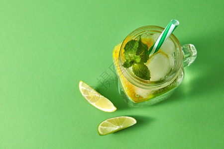 绿色桌上的玻璃泥瓦罐方有自制火花鸡尾酒冰石灰片柠檬绿色薄荷和塑料草冷酒精或非夏季饮料的概念用泥瓦罐中的冰石灰片和柠檬薄荷叶绿色背图片