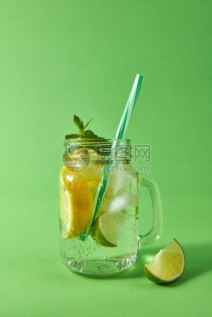 绿色桌上的玻璃罐子面有自制的火花鸡尾酒包括冰块石灰片和柠檬薄荷绿色叶子和塑料稻草冷酒精或非夏季饮料的概念图片