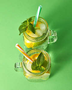 柠檬和冰薄荷叶玻璃上含塑料稻草的柑橘水果片绿色背景的两瓶玻璃罐绿色背景的柠檬和水玻璃上的塑料稻绿色背景的柠檬和水果片两瓶玻璃罐带图片
