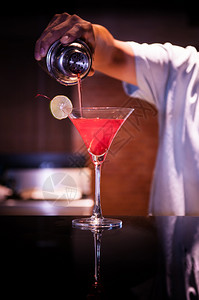 酒保把红马提尼鸡尾酒倒到一杯马丁尼子里面有石灰片酒吧里的暗光气图片