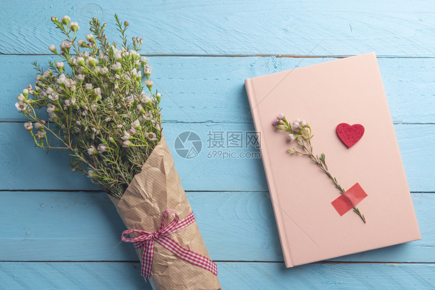 花束上面绑着红弓和粉色笔记本放在蓝色木制桌子上图片