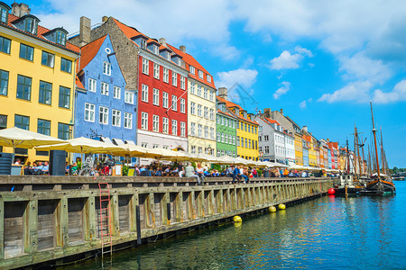 在阳光明媚的日子里在有咖啡馆和餐的Nyhavn河堤岸上旅游的客乘运河面甲根登山的船图片